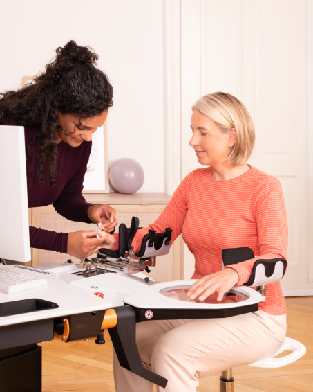 A woman is training with the Amadeo RMT module. A female therapist is sitting by her side.