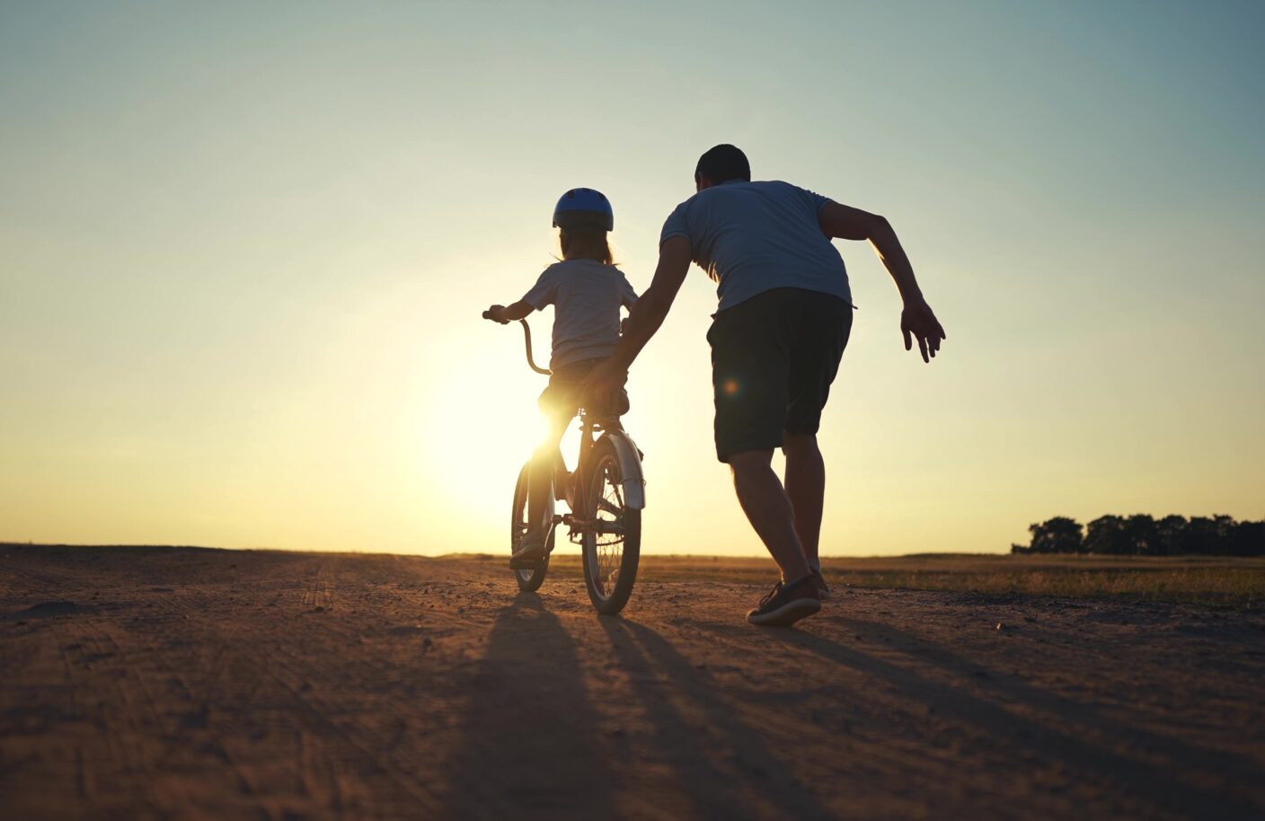 A child is riding a bicycle and a man is gently pushing the child. Both are seen from behind. The sun is shining.