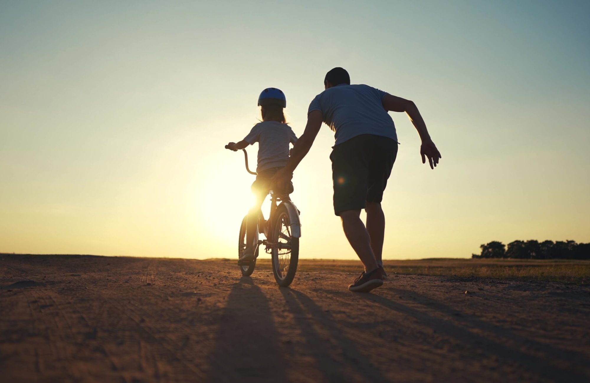 A child is riding a bicycle and a man is gently pushing the child. Both are seen from behind. The sun is shining.