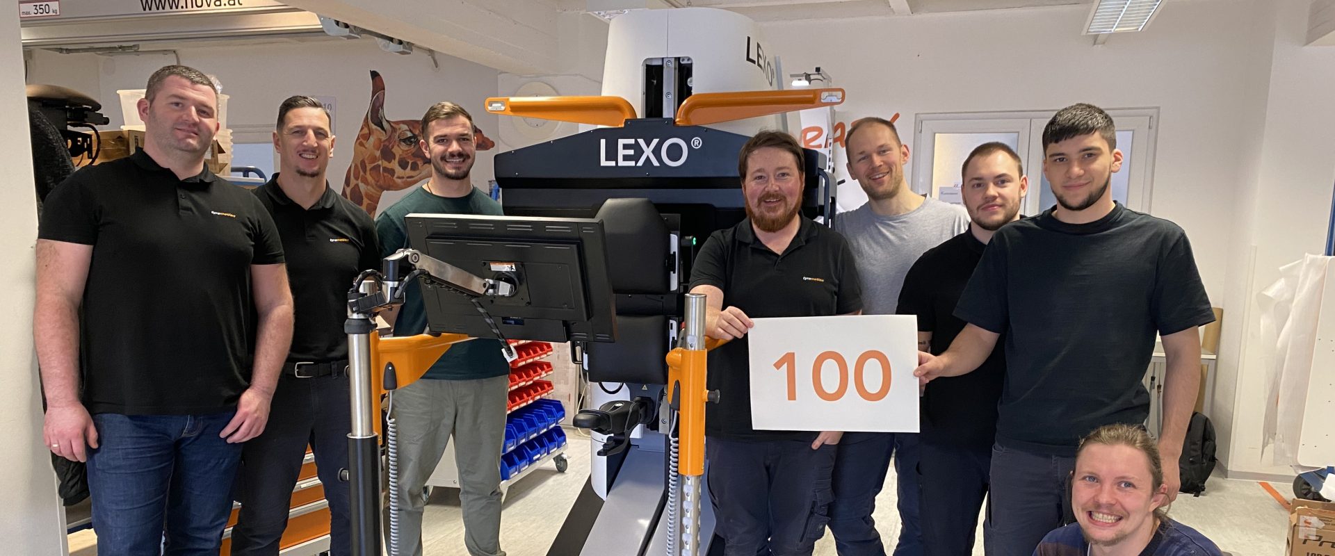 Picture of the Tyromotion Production Team in front of the gaittrainer Lexo and a sign with the number 100 on it