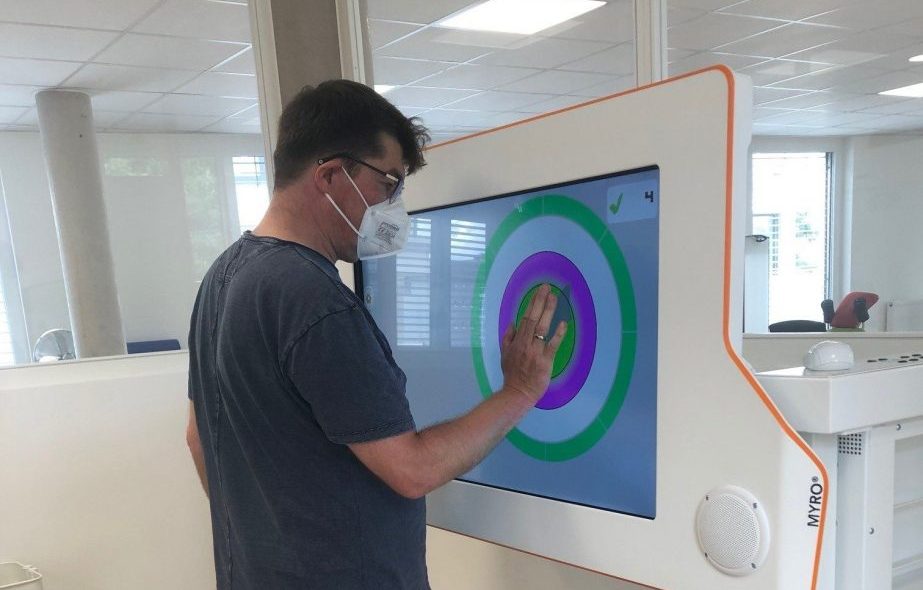Olaf wearing a face mask, standing in front of the Myro interactive rehabilitation device and holding his hand on it